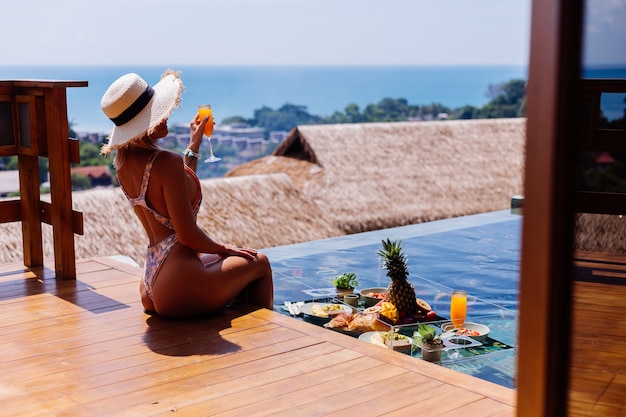 Belle femme bronzée caucasienne en bikini et chapeau de paille avec petit-déjeuner flottant à l'incroyable villa de style bali de luxe à la journée ensoleillée au bord de la piscine, fond tropical.
