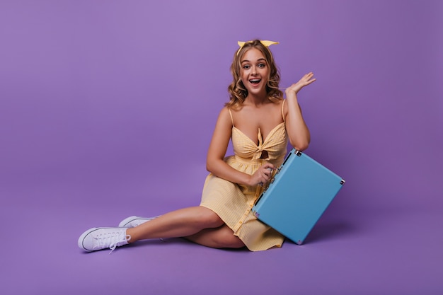 Belle femme bronzée avec sourire surpris drôle posant avec valise. Portrait de fille incroyable détendue en robe brillante.