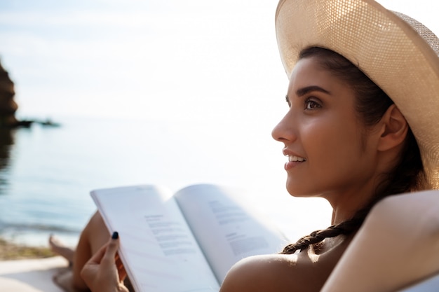 Belle femme brune au chapeau, lecture de livre, allongé sur la plage