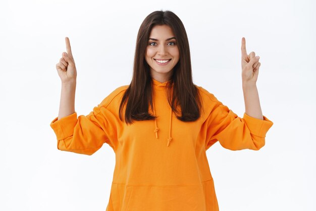 Belle femme brune confiante en sweat à capuche orange vous montrant le meilleur prix pointant les doigts vers le haut et regardant la caméra avec un sourire satisfait rayonnant annonce un produit présentant une nouvelle application