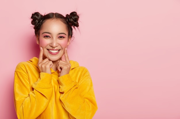 Belle femme brune avec deux petits pains, pointe l'index sur les joues, vêtue d'un sweat-shirt jaune en velours côtelé décontracté, porte un maquillage pin-up brillant