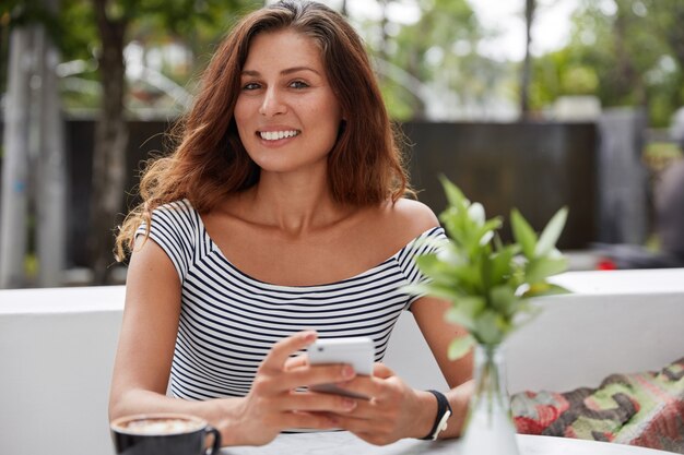 Belle femme brune avec une expression heureuse et téléphone dans un café terrasse en plein air