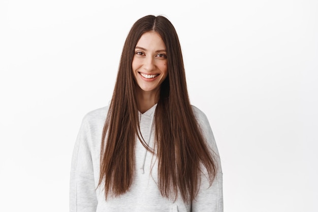 Belle femme brune avec de longs cheveux brillants en bonne santé souriant et regardant une publicité heureuse de soins capillaires et cosmétiques de beauté debout en sweat à capuche sur fond blanc