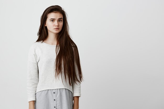 belle femme brune avec une longue coiffure posant contre le mur gris, gardant les lèvres entrouvertes