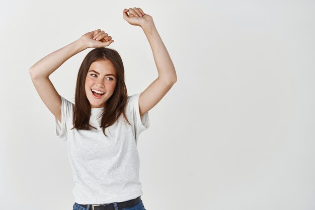 Belle femme brune s'amusant, dansant à l'intérieur avec les mains levées et souriante, debout heureuse sur un mur blanc.