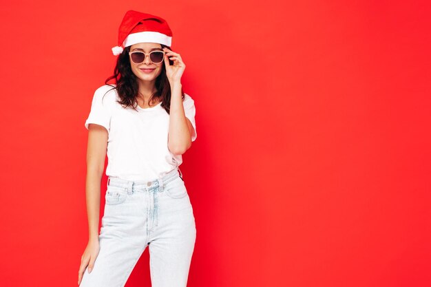 Belle femme célébrant le Nouvel An. Heureuse femme magnifique dans des vêtements décontractés s'amusant à la fête du Nouvel An. Célébration de vacances. Modèle charmant en chapeau de Père Noël riant près du mur rouge