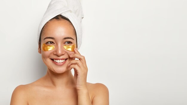 Belle femme charmante avec une peau saine, applique des patchs cosmétiques sous les yeux, regarde volontiers de côté, pense à quelque chose d'agréable, porte une serviette enveloppée sur la tête, a des procédures de spa à la maison