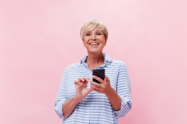 Belle femme en chemise à carreaux pose joyeusement sur fond rose et détient smartphone