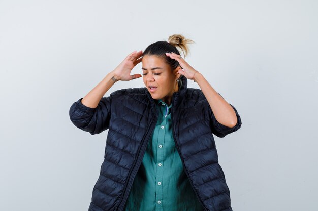 Belle femme en chemise verte, veste noire avec les mains sur les tempes et l'air ennuyé, vue de face.