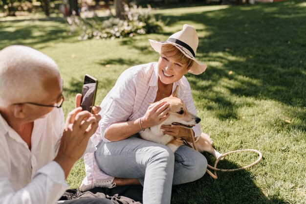 Belle femme avec une coiffure blonde cool en chapeau et chemise moderne rayée posant avec un chien et assis sur l'herbe avec un homme avec un téléphone en plein air.