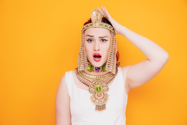 Belle femme comme Cléopâtre en costume égyptien antique confus et surpris tenant la main sur sa tête pour erreur sur orange