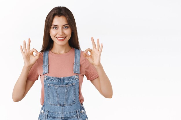 Belle femme confiante et affirmée en salopette en jean, t-shirt, montrant un signe d'approbation d'accord avec le sourire, d'accord ou d'accepter une bonne idée, debout sur fond blanc heureux, satisfait