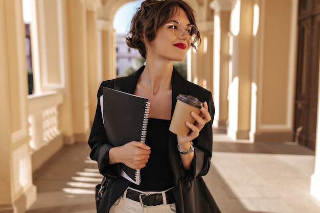 Photo gratuite belle femme dans des verres tenant une tasse de thé et un ordinateur portable à l'extérieur. femme brune aux lèvres rouges en veste noire souriant à l'extérieur.