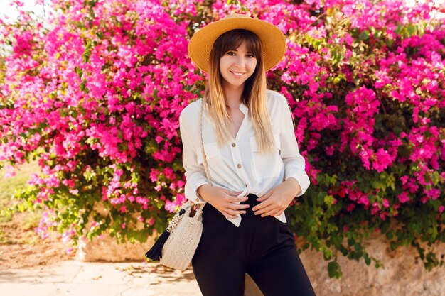 Belle femme debout sur des fleurs roses portant un chapeau de paille et une tenue décontractée.