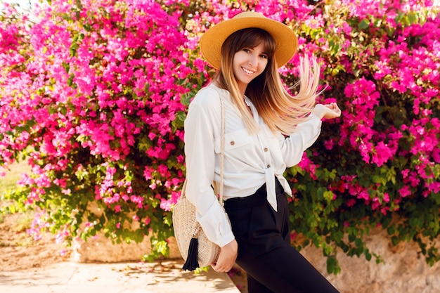 Belle femme debout sur des fleurs roses portant un chapeau de paille et une tenue décontractée.