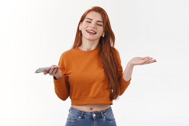 Une Belle Femme Détendue Et Insouciante écoute Toujours De La Musique Pendant Qu'elle Travaille, Porte Des écouteurs Sans Fil, Tient Un Smartphone Et écarte Les Mains Sur Le Côté, Ferme Les Yeux Et Profite D'un Bon Casque Audio