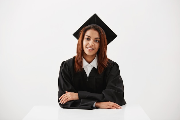 Belle femme diplômée souriant regardant la caméra assis sur une surface blanche