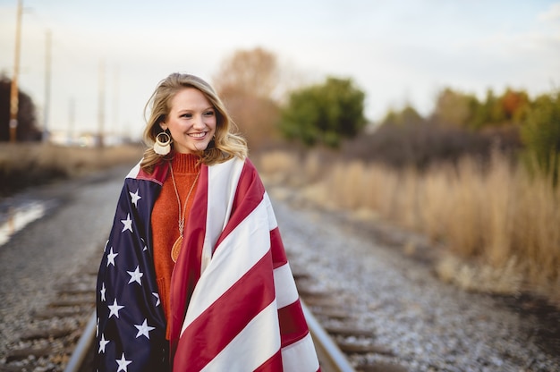 Belle femme avec le drapeau américain autour de ses épaules marchant sur le chemin de fer