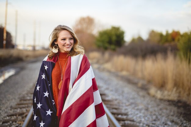 Belle femme avec le drapeau américain autour de ses épaules marchant sur le chemin de fer
