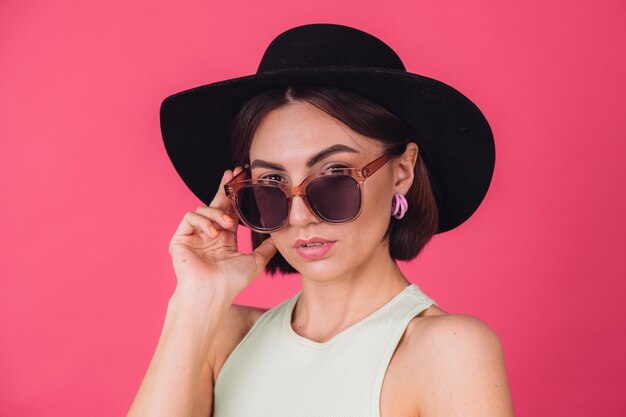 Belle femme élégante en chapeau et lunettes de soleil posant sur un mur rouge rose