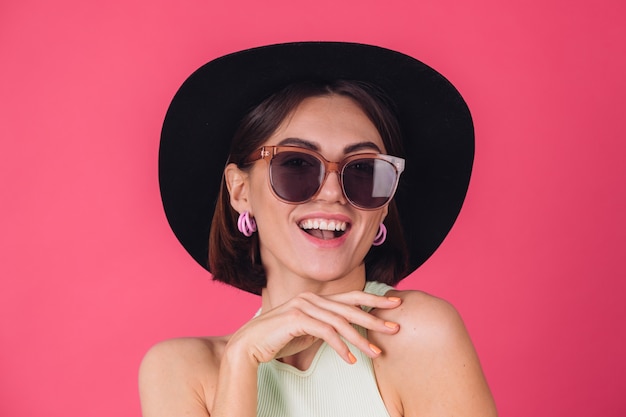 Belle femme élégante en chapeau et lunettes de soleil posant sur un mur rouge rose