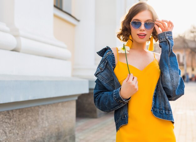 Belle femme élégante hipster posant, mode de la rue, tenant une fleur, robe jaune, veste en jean, style boho, tendance de la mode printemps été, sourire, lunettes de soleil bleues à la mode, souriant, ensoleillé