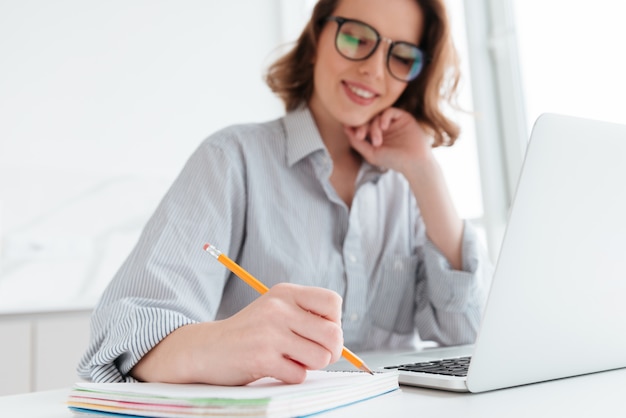 Belle femme élégante en vêtements décontractés, écrire des notes tout en étant assis dans une cuisine légère, mise au point sélective sur place