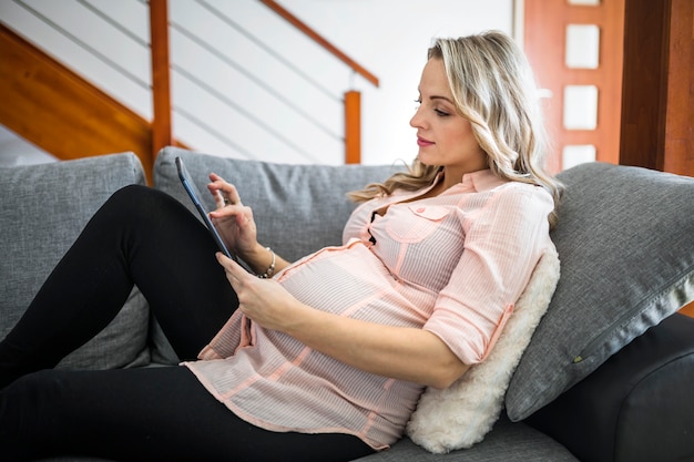 Belle femme enceinte à l'aide de tablette numérique sur canapé