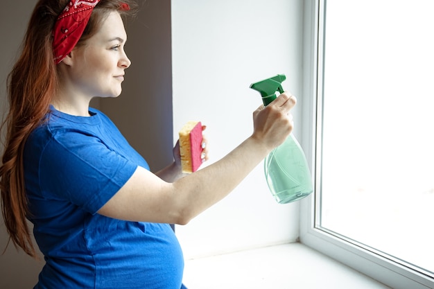 Une belle femme enceinte dans les derniers mois de la grossesse est engagée dans le nettoyage et lave les fenêtres.