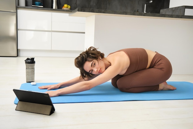 Photo gratuite belle femme européenne pratiquant le yoga de pleine conscience à la maison, assise sur un tapis de yoga, souriante et heureuse d'utiliser