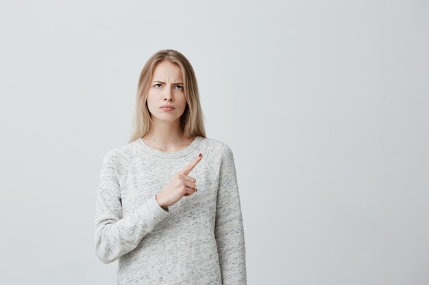 Belle femme furieuse aux cheveux teints blonds regardant de côté avec une expression sérieuse et en colère pointant l'index, montrant l'espace de copie