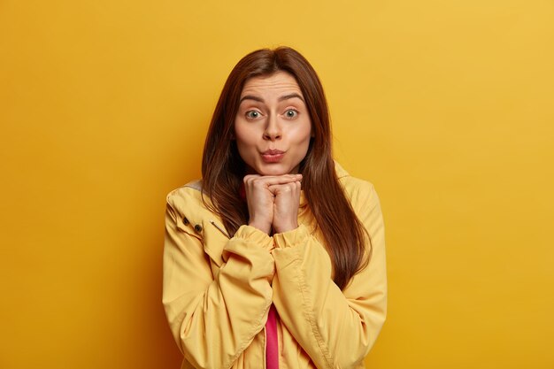 Belle femme garde les lèvres pliées, veut embrasser quelqu'un, regarde directement la caméra, tient les mains sous le menton, porte un anorak jaune