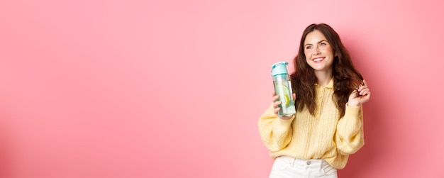 Belle femme glamour jouant avec les cheveux et souriant rêveur regardant de côté boire une boisson saine à partir d'une bouteille d'eau debout sur fond rose