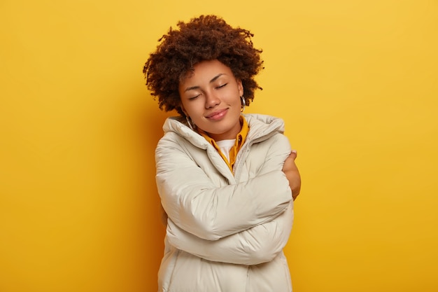 Photo gratuite belle femme heureuse apprécie le confort dans une nouvelle veste d'hiver, s'embrasse, garde les yeux fermés, se sent chaude et satisfaite, coiffure bouclée, isolée sur fond jaune. gens, concept de vêtements