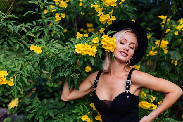 Belle femme heureuse caucasienne élégante en robe noire et chapeau classique dans le parc entouré de fleurs thaïlandaises jaunes