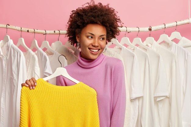 Belle femme heureuse choisit des vêtements en magasin, regarde volontiers de côté, tient un pull jaune sur des cintres