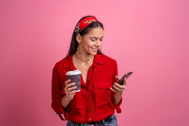 Belle femme hispanique en chemise rouge souriant tenant un smartphone buvant du café sur fond de studio rose isolé portant un bandeau