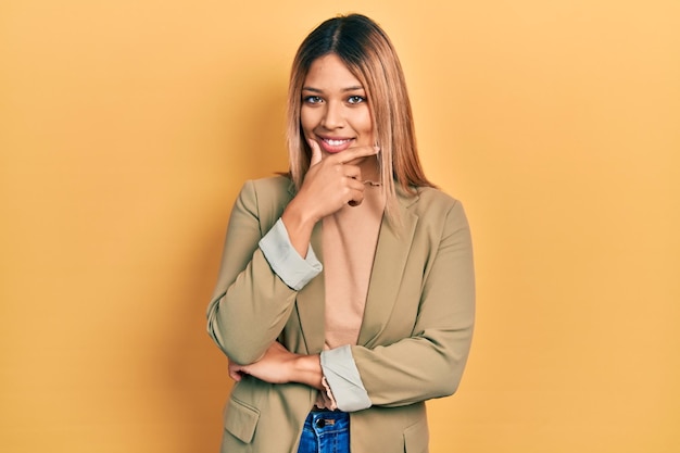 Belle femme hispanique portant une veste d'affaires à la recherche de confiance à la caméra en souriant avec les bras croisés et la main levée sur le menton en pensant positif