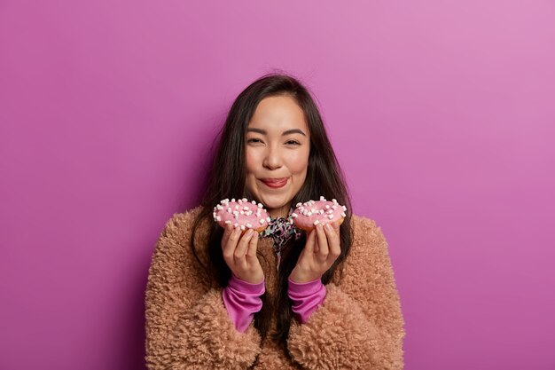 Belle femme lèche les lèvres avec la langue, tient deux beignets, a bon appétit, aime les produits sucrés, anticipe un goût agréable, a une expression heureuse