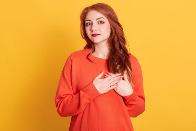 Belle femme avec un look amical, garde les mains sur la poitrine, exprime une attitude positive et bonne, a les cheveux ondulés rouges, ressent de la gratitude envers sa mère