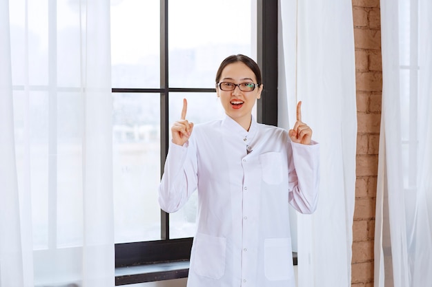 Belle femme avec des lunettes en blouse de laboratoire debout près de la fenêtre et pointant vers le haut.
