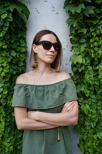 Belle femme en lunettes de soleil marron posant à côté des plantes. Photo extérieure de la magnifique jeune femme en tenue verte posant avec les mains croisées entre les feuilles vertes