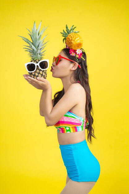 Belle femme en maillot de bain tenant un ananas pose sur jaune