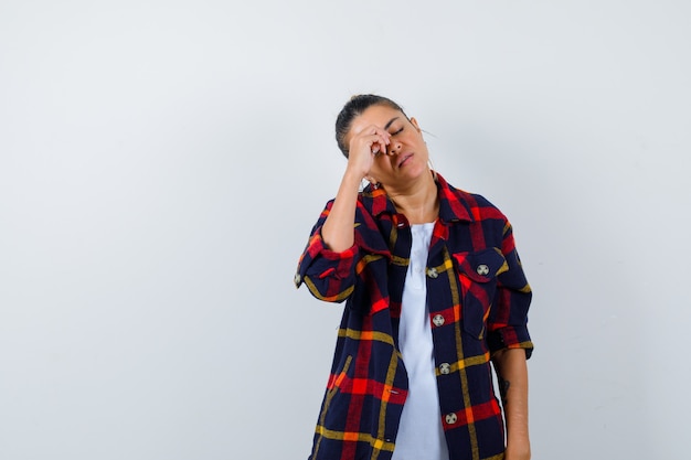 Belle femme avec la main sur les yeux en t-shirt blanc, chemise à carreaux et l'air fatigué, vue de face.