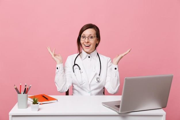 Belle femme médecin assise au bureau travaille sur ordinateur avec un document médical à l'hôpital isolé sur fond de mur rose pastel. femme en robe médicale lunettes stéthoscope. concept de médecine de santé