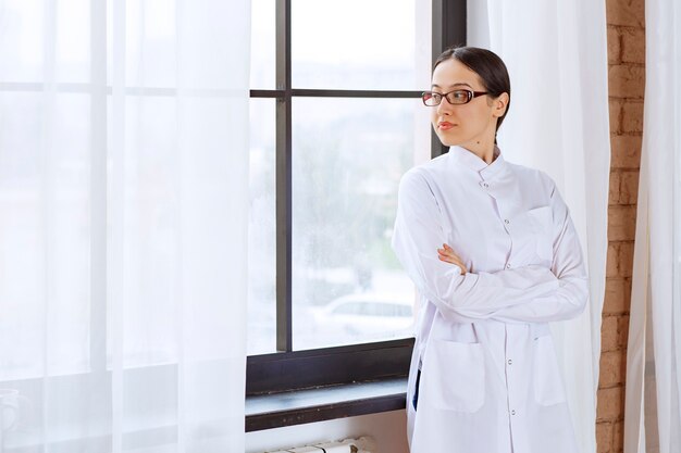 Belle femme médecin à lunettes debout près de la fenêtre et à la recherche.