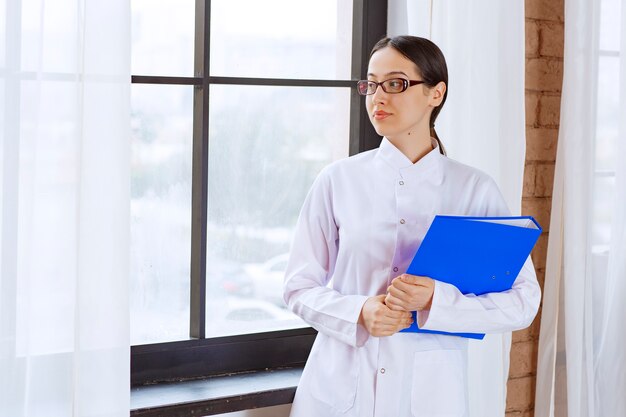 Belle femme médecin à lunettes avec dossier regardant quelque part près de la fenêtre.