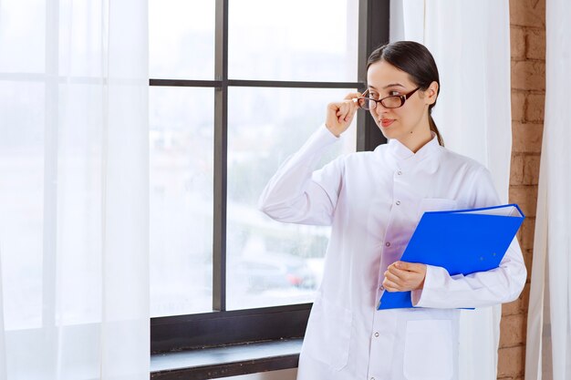 Belle femme médecin à lunettes avec dossier regardant quelque part près de la fenêtre.