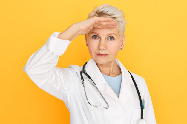 Belle femme médecin senior avec une coiffure courte teinte et des yeux bleus gardant la main sur son front à la recherche de quelque chose de loin à distance.