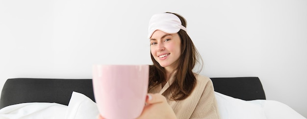 Photo gratuite belle femme moderne souriante en pyjama et masque de couchage offrant une tasse de thé ou de café au lit et